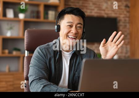 Souriant beau adulte asiatique homme avec casque agitant la main dans la webcam d'ordinateur, a des appels vidéo d'affaires Banque D'Images