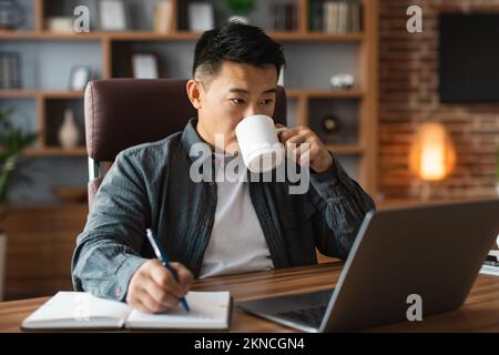 Un homme asiatique d'âge moyen très occupé boit du café, regarde l'ordinateur à table, a un appel vidéo à la maison Banque D'Images