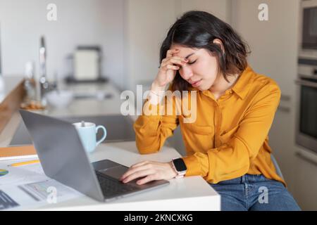 Femme indépendante arabe stressée souffrant de maux de tête en travaillant avec un ordinateur portable dans la cuisine Banque D'Images