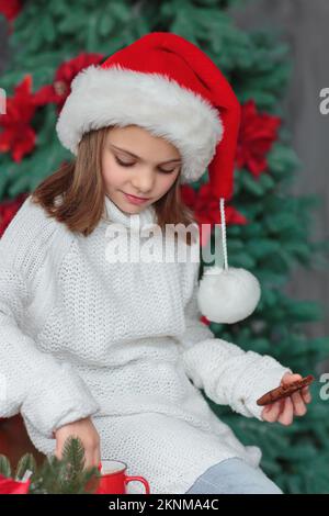 Enfant assis dans la cuisine confortable décorée de Noël et portant le chapeau rouge du père noël Banque D'Images