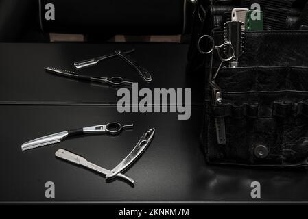 Outils de coiffure pour hommes, ciseaux de coupe de cheveux sur une surface noire lisse et un sac en cuir noir plein de séparateurs et de poches Banque D'Images
