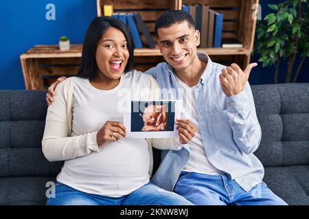 Un jeune couple hispanique attend un bébé assis sur le canapé montrant une échographie de bébé pointant le pouce vers le côté souriant heureux avec la bouche ouverte Banque D'Images