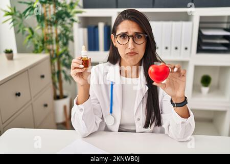 Jeune femme hispanique médecin tenant le coeur et l'huile de cbd déprimé et s'inquiéter de la détresse, pleurant en colère et peur. expression triste. Banque D'Images