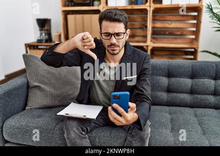 Jeune psychologue hispanique, homme en train de faire une thérapie par appel vidéo avec un smartphone au visage en colère, signe négatif montrant le détestant avec les pouces vers le bas, rejet Banque D'Images