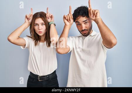 Jeune couple portant des vêtements décontractés debout ensemble faisant un geste drôle avec le doigt au-dessus de la tête que les cornes de taureau Banque D'Images