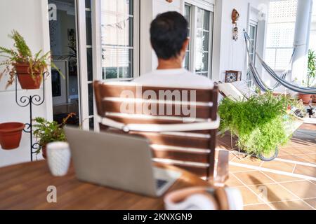 Homme d'âge moyen utilisant un ordinateur portable à la maison debout en arrière regardant loin avec les bras croisés Banque D'Images