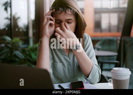 Une jeune femme fatiguée et stressée tient des lunettes et se moque de son nez tout en travaillant dur dans un café. Banque D'Images