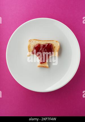 Pain grillé croustillant avec confiture de fraises et deux petites bouchées manquantes sur une assiette blanche et un fond rose vif Banque D'Images