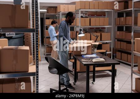 Le responsable de magasin prépare les commandes des clients en plaçant les produits dans des boîtes de carton, en vérifiant les détails d'expédition avant les colis de livraison. Équipe diversifiée travaillant dans l'entrepôt du centre de distribution Banque D'Images