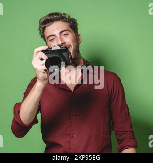 Un beau photographe branché qui passe en revue des photos de l'écran numérique d'un appareil photo sans miroir, prise en studio, isolé sur fond vert Banque D'Images