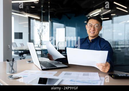 Portrait réussi satisfait asiatique comptable, homme en chemise et lunettes travaillant à l'intérieur du bureau à l'aide d'une calculatrice et d'un ordinateur portable pour les comptes et les rapports de documents financiers, regardant l'appareil photo. Banque D'Images