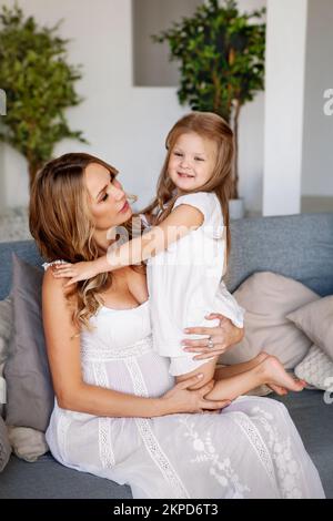 Portrait de la mère enceinte avec l'enfant passant du temps ensemble à la maison. Concept de style de vie maman et fille sont heureux. Famille assise sur un canapé, portant Banque D'Images