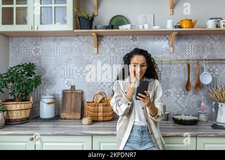 Une heureuse femme latino-américaine est debout dans la cuisine à la maison, tenant le téléphone d'une main, tenant sa tête de l'autre main. Je suis heureux, j'ai reçu un message inattendu et joyeux, de bonnes nouvelles. Banque D'Images