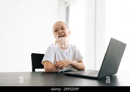 Rire blonde petite fille avec la main cassée enveloppée dans le bandage en plâtre blanc assis à la table avec ordinateur portable regardant joyeusement loin dans la lumière Banque D'Images