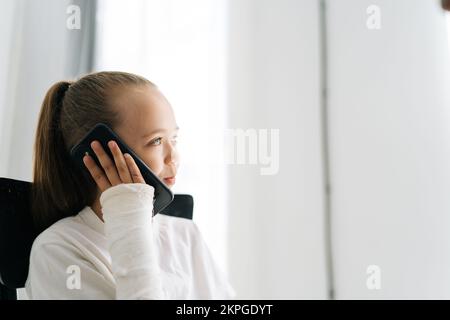 Vue latérale d'une petite fille blonde douce avec bras cassé enveloppé dans un bandage en plâtre parlant smartphone, souriant regardant loin sur fond blanc. Banque D'Images