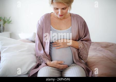 Une femme enceinte heureuse qui se couche le ventre, assise sur le lit. Banque D'Images