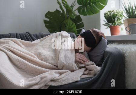 La jeune fille attirante dans le masque noir dormant pour les yeux dort ou nap sur le canapé dans la chambre. Concept de sieste de jour. Mise au point sélective Banque D'Images