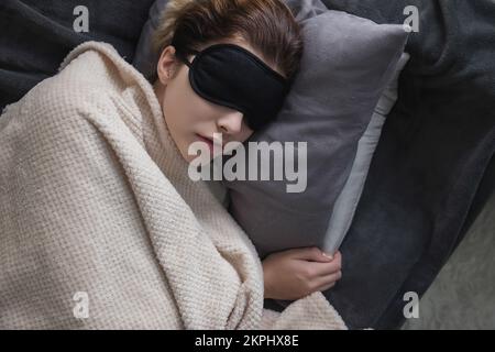 La jeune fille attirante dans le masque noir dormant pour les yeux dort ou nap sur le canapé dans la chambre. Concept de sieste de jour. Mise au point sélective Banque D'Images