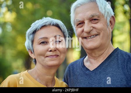 Un vieux couple optimiste et actif de 60s ans en amour pose à l'extérieur, sourire en regardant la caméra pendant une promenade matinale dans un parc public. Joyeux mariage sans fin, romantis Banque D'Images