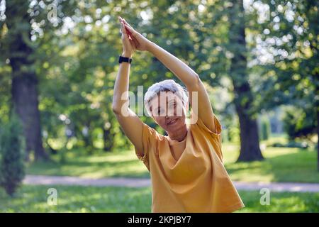 Femme asiatique active plus âgée dans les vêtements de sport regarde la caméra méditant dans le parc d'été avec des paumes repliées fait des exercices d'étirement, faire de l'exercice de yoga Banque D'Images