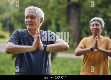 Couple actif plus âgé pratiquer la méditation avec les yeux fermés poser dans le parc d'été bras pliés fait mudra ou signe Namaste, faire l'exercice de yoga dehors, lui Banque D'Images
