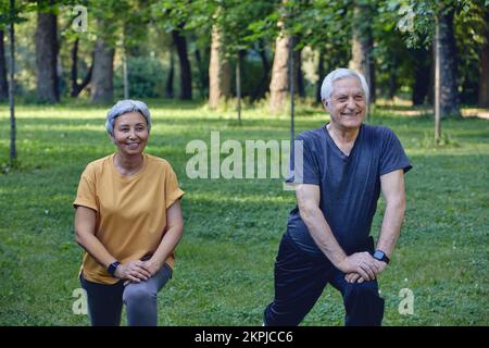 Les conjoints âgés font des exercices sportifs à l'extérieur dans le parc d'été le matin. Un couple plus âgé en bonne santé, en train de se réchauffer, en faisant des squats sourire, profitez d'une activité active Banque D'Images