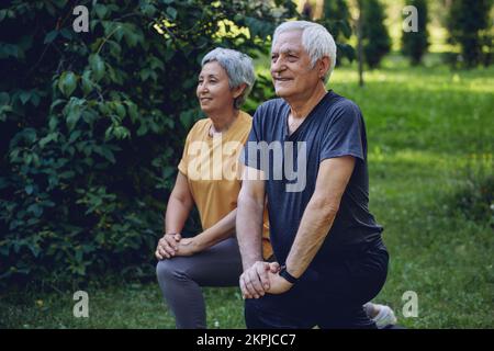 Les conjoints âgés font des exercices sportifs à l'extérieur dans le parc d'été le matin. Un couple plus âgé en bonne santé, en train de se réchauffer, en faisant des squats sourire, profitez d'une activité active Banque D'Images