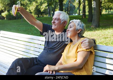 Des conjoints heureux prennent photo de selfie assis sur le banc dans le parc d'été. Couple gris-cheveux regarder l'écran de téléphone cellulaire faire videocall utilisation extérieure moderne t Banque D'Images
