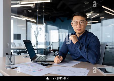 Portrait d'un investisseur asiatique sérieux et mature, homme d'affaires en lunettes et chemise regardant attentivement et concentré à la caméra, homme travaillant avec des documents factures et rapports à l'intérieur du bureau. Banque D'Images