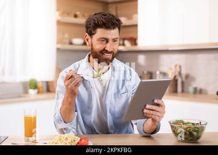 Un homme de race blanche, content d'être un adulte, mange un déjeuner, lit les actualités sur une tablette, regarde la vidéo dans un intérieur de cuisine moderne Banque D'Images