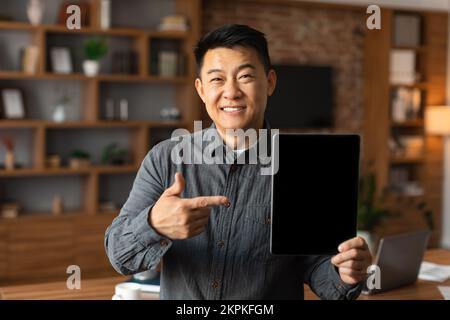 Souriant homme chinois d'âge moyen pdg montrer le doigt sur une tablette avec écran vide à l'intérieur du bureau à domicile Banque D'Images