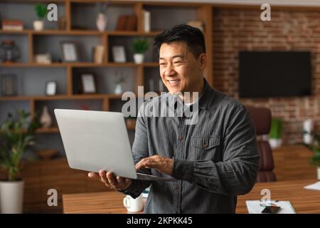 Souriant beau, homme asiatique d'âge moyen directeur général dactylographiant sur ordinateur portable, a appel vidéo à l'intérieur du bureau à domicile Banque D'Images