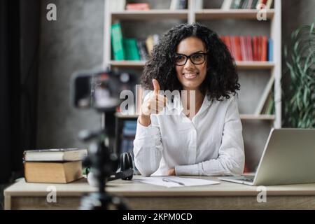 Souriante femme multiraciale en chemise blanche assise au bureau avec des livres, des notes et un ordinateur portable enregistrant un blog vidéo et montrant les pouces vers le haut. Femme enseignant travaillant à la maison pendant l'apprentissage à distance. Banque D'Images