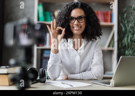 Femme souriante multiraciale en chemise blanche assise à un bureau avec des livres, des notes et un ordinateur portable enregistrement vidéo blog et montrant signe ok. Femme enseignant travaillant à la maison pendant l'apprentissage à distance. Banque D'Images