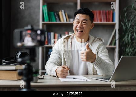 Homme asiatique souriant dans des vêtements décontractés assis au bureau avec des livres, des notes et un ordinateur portable enregistrant un blog vidéo et montrant le pouce vers le haut. Tuteur masculin travaillant à la maison pendant l'apprentissage à distance. Banque D'Images