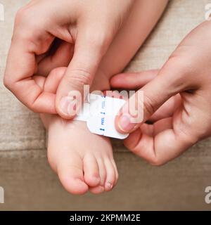 La mère colle un pansement médical adhésif sur la jambe du bébé. Main de maman avec ruban adhésif de protection collant et pied d'enfant. Enfant âgé de un vous Banque D'Images