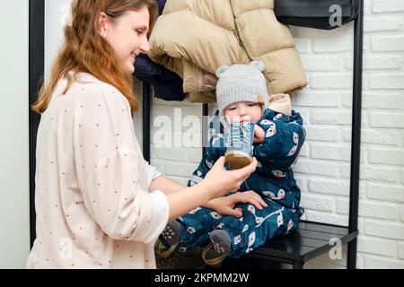 La mère met des chaussures bleues sur le bébé assis dans le couloir de la maison. Femme maman portant chaud bottes vêtements sur enfant pour l'hiver marcher dans le froid W Banque D'Images