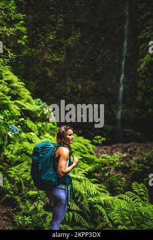 Description: Femme sportive se présente dans la nature à une impressionnante cascade dans la jungle avec de belles fleurs et fougères. Levada de Caldeirão Verde, fabriqué Banque D'Images