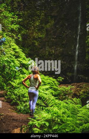Description: Femme sportive se présente à une impressionnante cascade dans la jungle avec de belles fleurs et fougères. Levada de Caldeirão Verde, île de Madère Banque D'Images