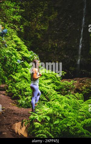Description: Femme sportive se présente dans la nature à une impressionnante cascade dans la jungle avec de belles fleurs et fougères. Levada de Caldeirão Verde, fabriqué Banque D'Images
