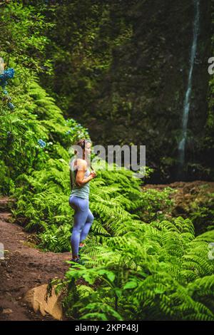 Description: Femme sportive se présente dans la nature à une impressionnante cascade dans la jungle avec de belles fleurs et fougères. Levada de Caldeirão Verde, fabriqué Banque D'Images