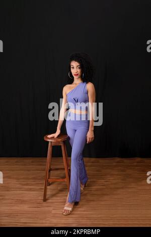 Portrait d'une jeune femme belle et joyeuse debout en studio à côté d'un tabouret. porter des vêtements bleus. Sur fond noir. Banque D'Images