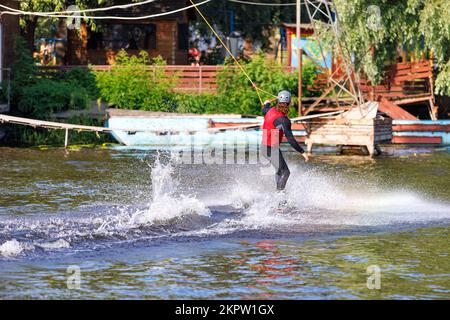 Un wakeboarder d'athlète coupe la surface de l'eau à grande vitesse sur un fond flou de la rive de la rivière créant un nuage de éclaboussures autour de lui. Banque D'Images