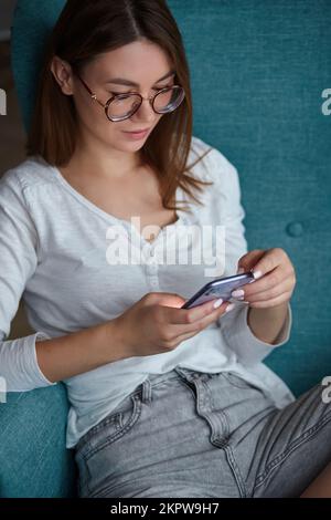Jeune femme heureuse utilisant un téléphone portable tenant dans les mains tout en étant assise dans un fauteuil à la maison Banque D'Images