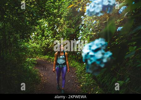 Description: Femme sportive avec sac à dos en admirant de belles fleurs sur le chemin vert de la jungle le long du canal d'eau. Levada de Caldeirão Verde, île de Madère Banque D'Images