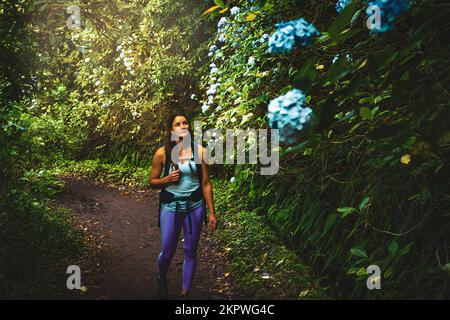 Description: Femme sportive avec sac à dos en admirant de belles fleurs sur le chemin vert de la jungle le long du canal d'eau. Levada de Caldeirão Verde, île de Madère Banque D'Images