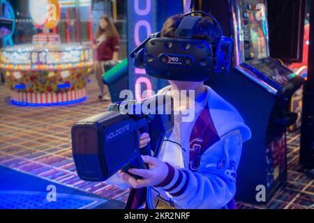 Un garçon jouant à un jeu vidéo de réalité virtuelle dans une arcade, tenant un fusil et portant un casque VR Banque D'Images