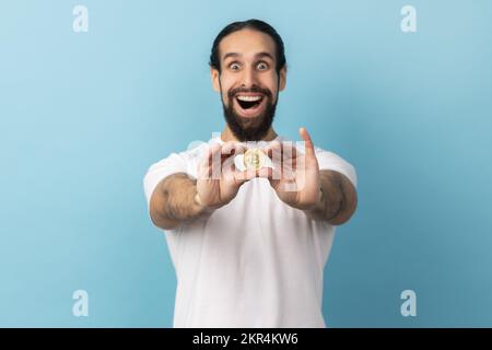 Portrait d'un homme surpris avec une barbe portant un T-shirt blanc tenant le bitcoin, prêtant attention à la nouvelle crypto-monnaie numérique. Studio d'intérieur isolé sur fond bleu. Banque D'Images