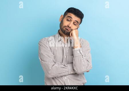 Portrait d'un homme d'affaires barbu triste ou ennuyé debout et regardant l'appareil photo avec un visage de tristesse insatisfait, portant une chemise rayée. Studio d'intérieur isolé sur fond bleu. Banque D'Images