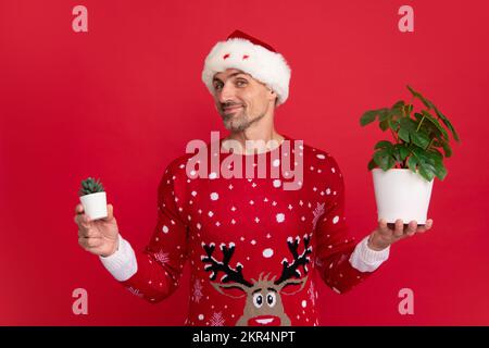 Homme en chandail de Noël et chapeau sur fond de couleur. Pot de conservation de Santa avec plante. père noël d'âge moyen. Banque D'Images